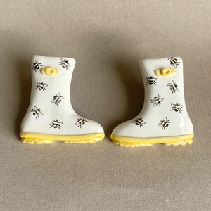 Peut inclure: Une paire de bottes de pluie en céramique blanche avec une bordure jaune et un motif d'abeilles.