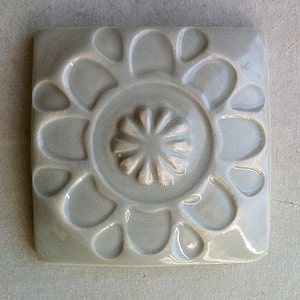 May include: A square ceramic tile with a raised, embossed design of a flower with a central circle. The tile is a light gray color.