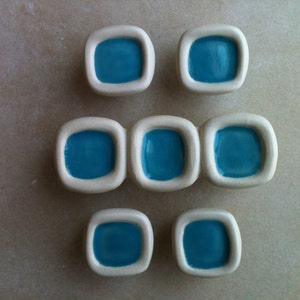 Peut inclure: Ensemble de huit carreaux de céramique carrés aux bords arrondis. Chaque carreau présente un centre turquoise vif entouré d'une bordure blanche. Les carreaux sont disposés en grille sur une surface de ton neutre.