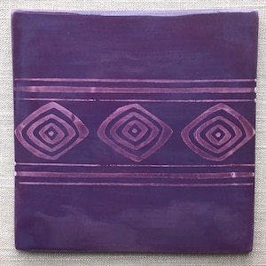 May include: A purple ceramic tile with a decorative pattern of three concentric squares in a diamond shape. The pattern is repeated three times across the tile and is outlined in a lighter shade of purple.