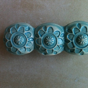 May include: Three teal blue ceramic tiles with a raised, circular, floral design. Each tile has a smaller, raised circle in the center.