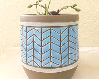Könnte beinhalten: Ein brauner Keramiktopf mit einem blauen und weißen Chevron-Muster. Der Topf ist mit einer kleinen Sukkulentenpflanze gefüllt.