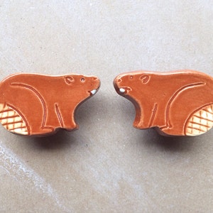 May include: Two ceramic brown beaver figurines with white accents. The beavers are facing each other and have a textured tail.