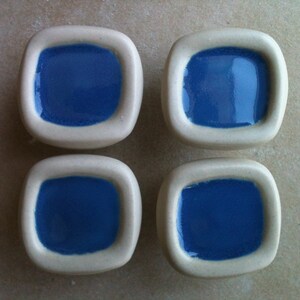 Könnte beinhalten: Vier quadratische Keramik-Schrankknöpfe mit weißem Rand und blauem Zentrum.