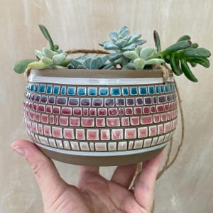May include: A hanging planter made of brown ceramic with a mosaic design in shades of pink, blue, and white. The planter is filled with succulents.