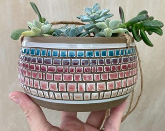 May include: A hanging planter made of brown ceramic with a mosaic design in shades of pink, blue, and white. The planter is filled with succulents.
