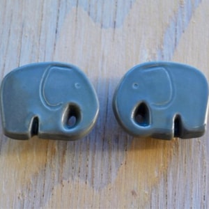 May include: Two gray ceramic elephant-shaped cabinet knobs. Each knob features a detailed elephant design with a trunk, ear, and a hole for the screw. These knobs are a decorative accent for furniture.
