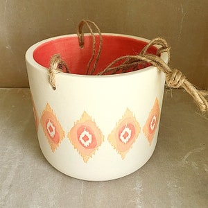 May include: White ceramic hanging planter with a pink and orange geometric pattern. The planter has a red interior and a jute rope for hanging.