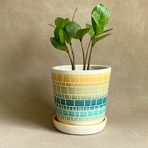 Puede incluir: Una planta verde en una maceta de cerámica con un diseño de mosaico amarillo, verde y azul. La maceta tiene un platillo a juego.