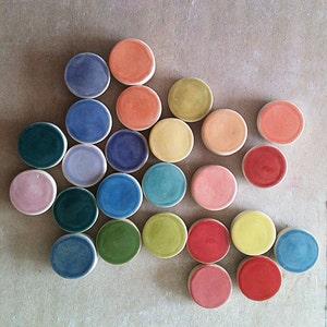 May include: A collection of 24 round ceramic tiles in various colors, including pink, red, orange, yellow, green, blue, and purple. The tiles are arranged in a semi-circular pattern.