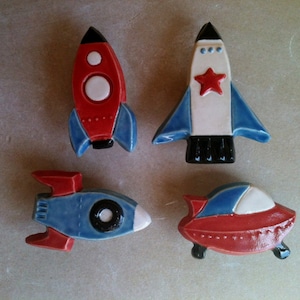 May include: Four ceramic rocket ship ornaments in red, blue, white and black. The ornaments are in the shape of rockets with different designs.
