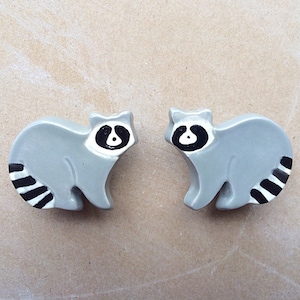 May include: Two ceramic raccoon figurines with black and white striped tails. The raccoons are gray with black eyes and black noses.