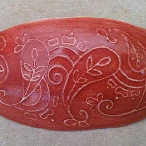 Può includere: Un piatto in ceramica rosso con un motivo paisley bianco. Il piatto è di forma ovale e ha una finitura lucida.