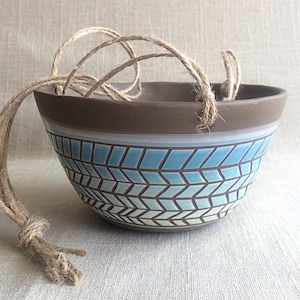 May include: A brown ceramic hanging planter with a blue and white geometric pattern. The planter has a rope handle.