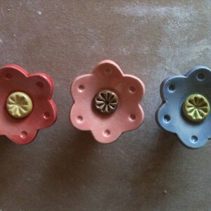 May include: Three ceramic flower-shaped knobs in pink, light pink, and blue with yellow centres. The knobs have a raised, textured design.