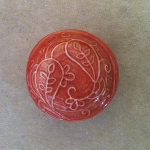 Può includere: Una piccola ciotola rotonda in ceramica arancione con un motivo floreale bianco. La ciotola è decorata con un motivo paisley e foglie.