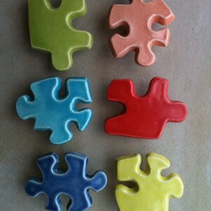 Pode incluir: Seis peças de quebra-cabeça de cerâmica coloridas em verde, pêssego, azul, vermelho, marinho e amarelo. Cada peça tem um acabamento brilhante e está disposta sobre uma superfície neutra. As peças são projetadas para se interligarem, sugerindo um jogo ou um item decorativo.