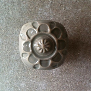 May include: A grey ceramic knob with a raised, circular design. The knob has a textured surface and a raised centre.