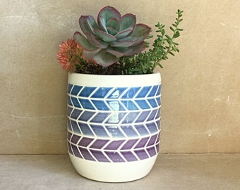 May include: A white ceramic planter with a geometric pattern in shades of blue and purple. The planter is filled with a succulent plant and a small green plant.