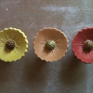 May include: Three ceramic flower-shaped knobs in yellow, peach, and red. Each knob has a raised centre with a brown, green, and brown centre, respectively.