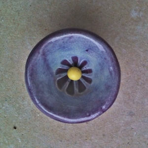 May include: A purple ceramic dish with a flower design and a yellow centre. The dish is glazed and has a slight sheen.