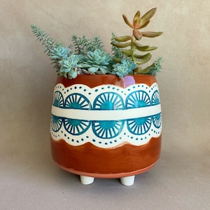May include: Terracotta planter with a white and teal geometric design. The planter has three small feet and is filled with succulents.