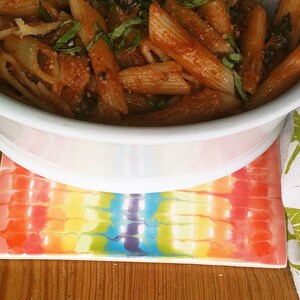 May include: A white bowl filled with penne pasta in a tomato sauce with fresh basil leaves. The bowl is sitting on a colorful rainbow-patterned coaster.