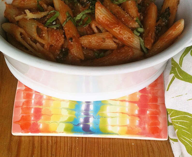 May include: A white bowl filled with penne pasta in a tomato sauce with fresh basil leaves. The bowl is sitting on a colorful rainbow-patterned coaster.