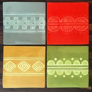 May include: Four square ceramic tiles with different colored backgrounds and white geometric designs. The tiles are arranged in a grid pattern.