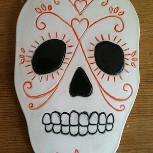 May include: White ceramic sugar skull with orange painted details. The skull has black eyes, a black nose, and a black mouth with teeth.