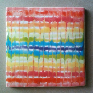 May include: A square ceramic tile with a colorful tie-dye pattern. The tile features a rainbow of colors, including red, orange, yellow, green, blue, and purple. The colors are arranged in horizontal stripes, with a white background.