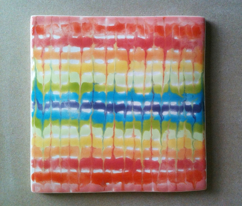 May include: A square ceramic tile with a colorful tie-dye pattern. The tile features a rainbow of colors, including red, orange, yellow, green, blue, and purple. The colors are arranged in horizontal stripes, with a white background.