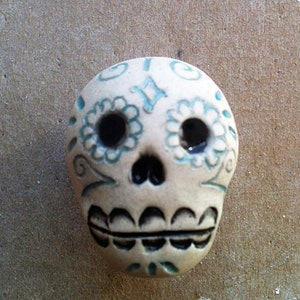 May include: White ceramic sugar skull with blue and black painted details. The skull has a traditional Day of the Dead design with floral patterns and a black mouth.