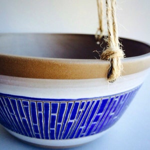 May include: A white ceramic hanging planter with a blue geometric pattern and a brown rim. The planter has a rope handle.