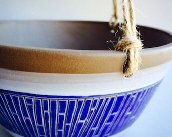 May include: A white ceramic hanging planter with a blue geometric pattern and a brown rim. The planter has a rope handle.