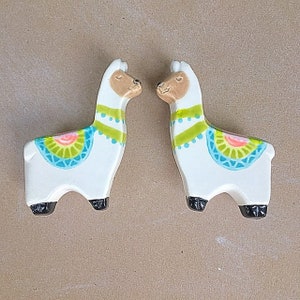 May include: Two ceramic llama salt and pepper shakers with white bodies and colorful geometric designs. The llamas are facing each other.