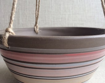 May include: A brown ceramic hanging planter with a striped design in shades of pink, beige, and white. The planter has a rope hanger.