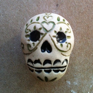 May include: A white ceramic sugar skull with green and black painted details. The skull has a heart shape on the forehead and a floral design around the eyes.