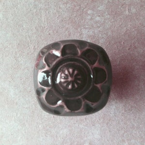 May include: A dark gray ceramic cabinet knob with a raised floral design. The knob has a circular center with a starburst pattern.