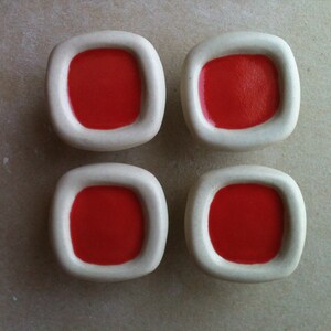 Peut inclure: Quatre boutons en céramique blanche de forme carrée avec un centre rouge. Les boutons ont un bord légèrement surélevé autour du centre rouge.