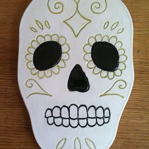 May include: White ceramic sugar skull with black eyes and a black mouth. The skull is decorated with green swirls and floral designs.