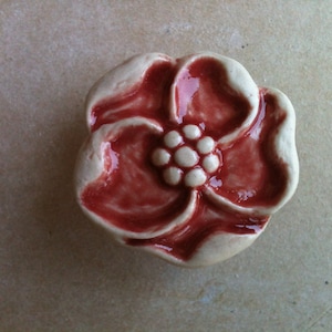 May include: A ceramic flower-shaped knob with a red glaze and a white centre. The knob is a decorative element for furniture or cabinets.