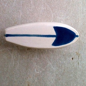May include: A white ceramic surfboard with a blue stripe and a blue arrow shape on the top.