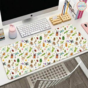May include: A rectangular desk mat with a cream background and a colorful pattern of insects, butterflies, and leaves. The mat is placed on a white desk, with a computer, keyboard, and other office supplies visible.