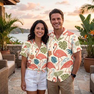 May include: Two short-sleeved button-down shirts featuring a cream background with a repeating pattern of pink flamingos, green tropical leaves, and orange hibiscus flowers. The shirts have a classic collar and a button-up front closure.