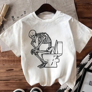 May include: White t-shirt featuring a black and white graphic of a skeleton sitting on a toilet, chin in hand, in a thoughtful pose. The shirt has a crew neck and short sleeves. The design is centered on the front of the shirt.