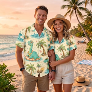 May include: A short-sleeved button-down shirt featuring a cream background with a repeating pattern of teal dinosaurs, palm trees, ocean waves, and small stars. The shirt has a notched collar and a button-front closure, showcasing a tropical theme.