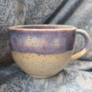 Könnte beinhalten: Eine handgefertigte Keramiktasse mit runder Form und großem Henkel. Die obere Hälfte hat einen Farbverlauf von Lila und Blau, während die untere Hälfte gesprenkeltes Creme ist. Die Tasse ist für Heißgetränke konzipiert.