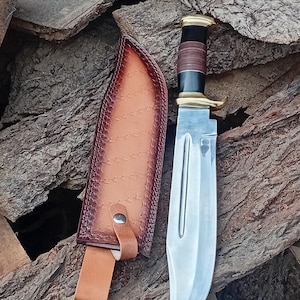 May include: A large hunting knife with a polished steel blade and a brown leather sheath. The handle features a black and brown grip with brass accents. The sheath is tan leather with decorative stitching and a belt loop.