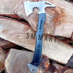 May include: A silver and black tactical axe with a toothed blade and a pointed end. The axe features a black handle with a textured grip and the text "SM MART" printed on the side. The axe is shown on a background of wooden logs.